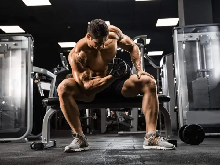 Mężczyzna z wyrzeźbionymi mięśniami wykonuje ćwiczenie na biceps z hantlem, skupiony na swoim **full body workout** w siłowni.