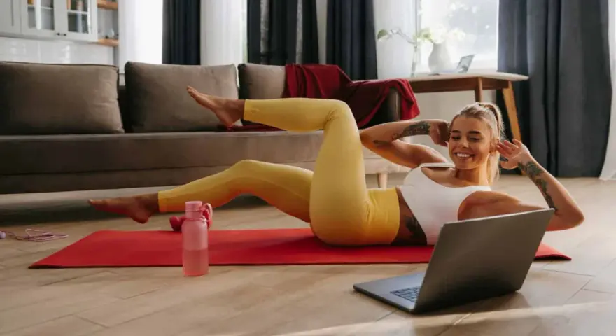 Uśmiechnięta kobieta wykonuje proste ćwiczenia w domu, leżąc na macie i patrząc na laptopa.