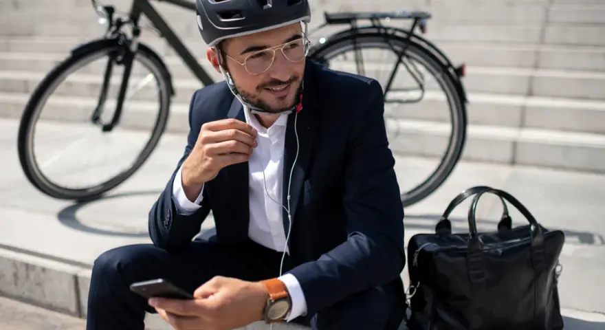 Mężczyzna w garniturze i kasku siedzi na schodach z rowerem w tle, słuchając muzyki przez słuchawki i przeglądając telefon.
