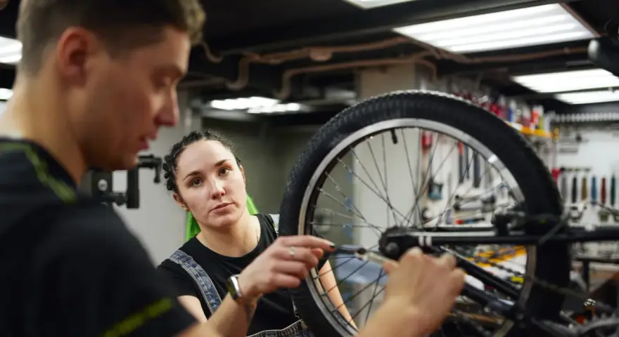 Mechanik rowerowy wyjaśnia, ile kosztuje wymiana dętki w rowerze, podczas gdy kobieta z zielonymi włosami uważnie słucha.