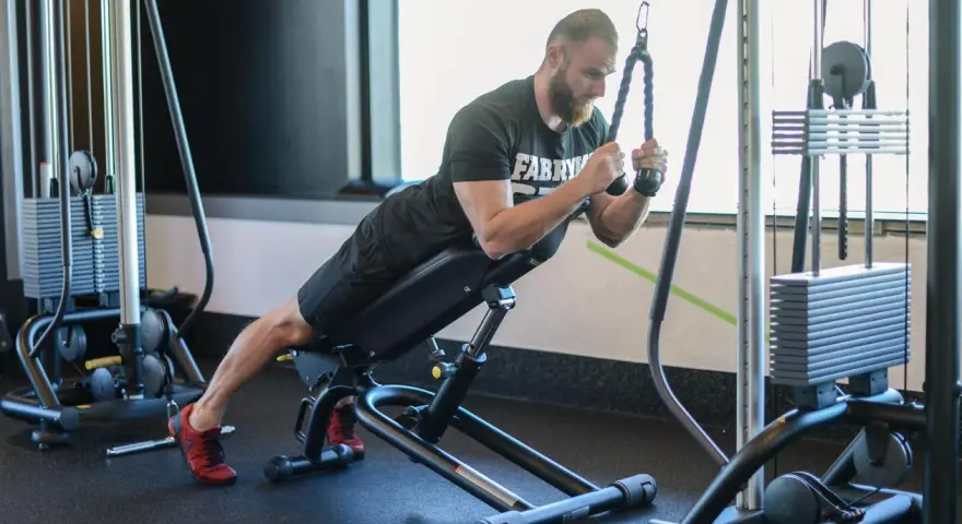 Mężczyzna wykonuje ćwiczenie na triceps, znane jako skull crushers, na ławce skośnej z użyciem wyciągu.