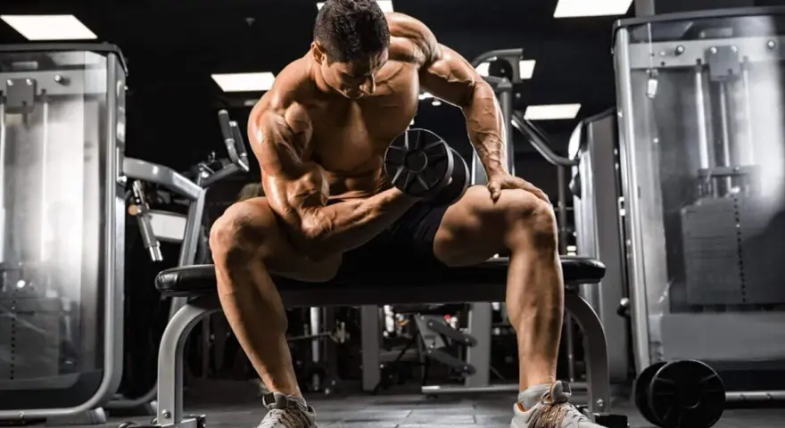 Mężczyzna z wyrzeźbionymi mięśniami wykonuje ćwiczenie na biceps z hantlem, skupiony na swoim **full body workout** w siłowni.