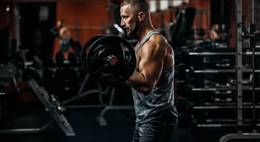 Mężczyzna ćwiczy biceps z ciężarem. W tle siłownia, sprzęt i inny ćwiczący.