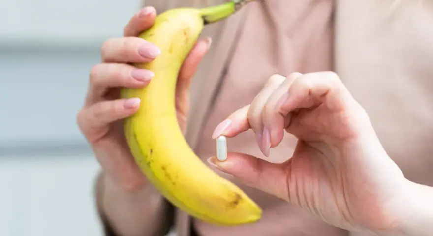 Kobieta trzyma banana i tabletkę, zastanawiając się, ile potasu dziennie powinna przyjmować.