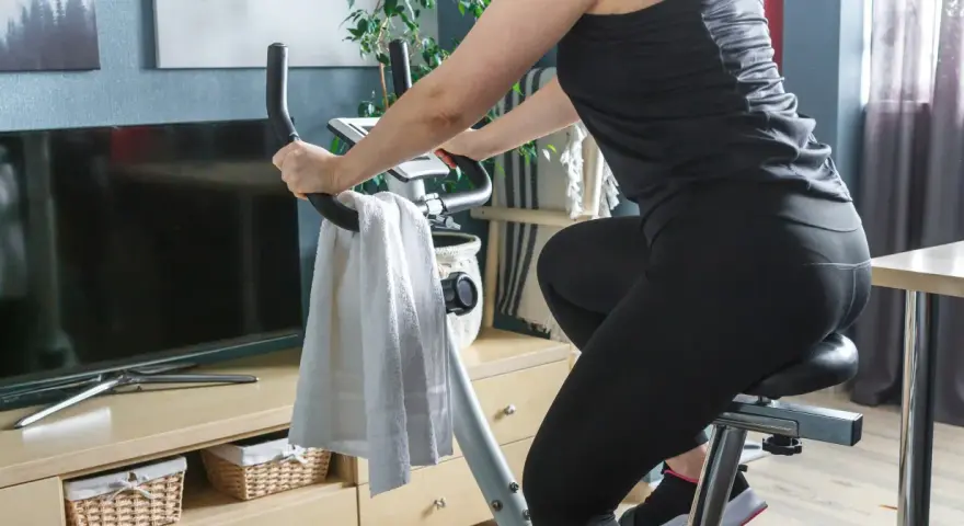 Kobieta ćwiczy na rowerku stacjonarnym, osiągając efekty w domowym zaciszu. Obok ręcznik i telewizor.