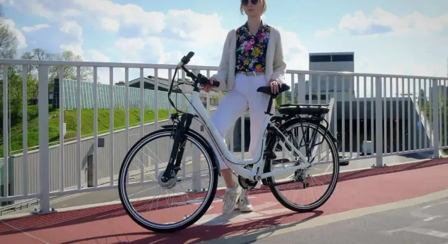 Kobieta w białych spodniach i kolorowej koszuli stoi obok białego roweru elektrycznego. Czy na rower elektryczny trzeba mieć prawo jazdy?