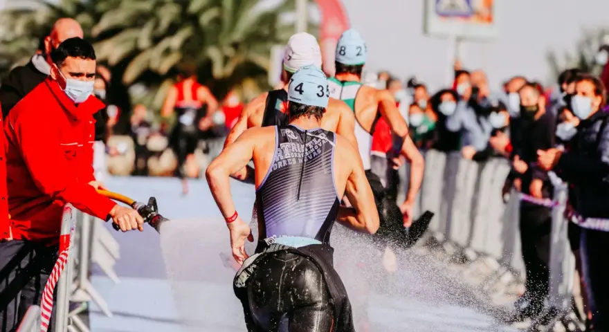 Zawodnicy triathlonu olimpijskiego biegną, a strażak polewa ich wodą z węża, by ich ochłodzić. Widownia w maskach obserwuje zza barierek.