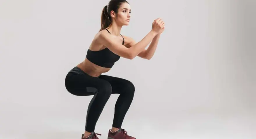 Kobieta w czarnym sportowym stroju wykonuje ćwiczenie air squats, z rękami splecionymi przed sobą.