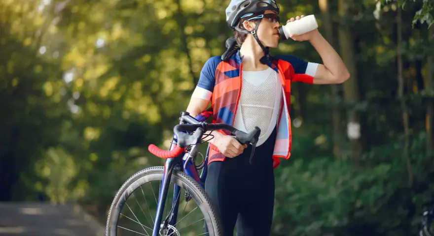 Kolarz w kasku i stroju sportowym pije wodę, trzymając rower. Dba o nawodnienie i dostarczenie makroskładników po wysiłku.