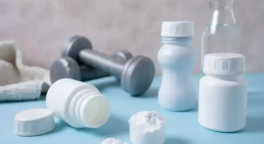 Białe pojemniki, miarka z proszkiem i hantle sugerują suplementację. Proteiny co to? To budulec mięśni, ważny dla sportowców.
