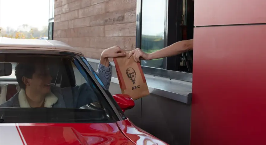 Mężczyzna w czerwonym aucie odbiera z okienka KFC torbę z jedzeniem. Wygląda na zadowolonego, że otrzymał swój mega pocket kcal.