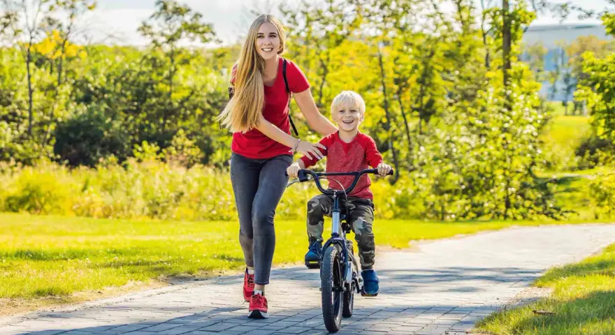 Mama uczy synka, jak nauczyć dziecko jeździć na rowerze. Radosny chłopiec pedałuje, a mama go asekuruje.