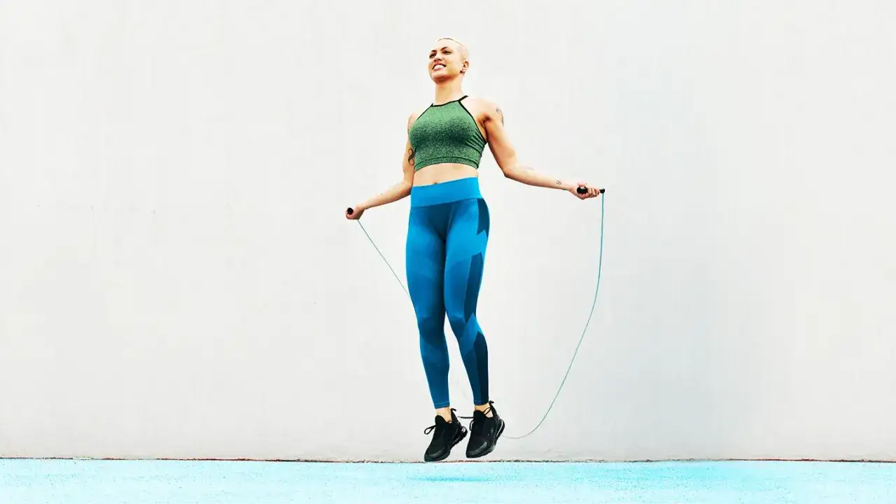 Uśmiechnięta osoba z krótkimi włosami skacze na skakance, ubrana w sportowy top i niebieskie legginsy.