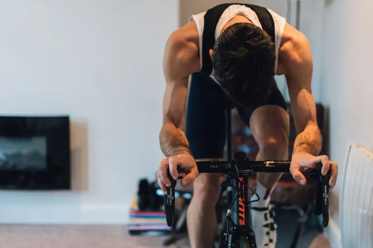 Intensywny trening HIIT na rowerze stacjonarnym. Mężczyzna w stroju kolarskim pochyla się nad kierownicą, widać jego spięte mięśnie.