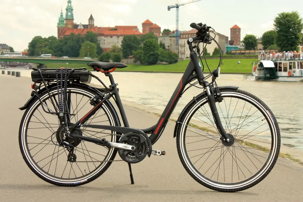 Czarny rower z bagażnikiem i baterią, gotowy do przerobienia na elektryczny. W tle Wawel i Wisła.