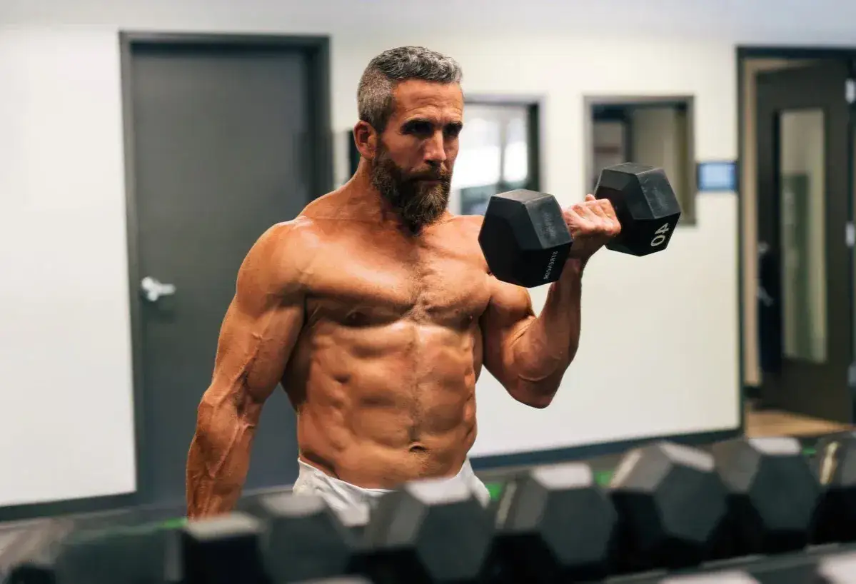 Mężczyzna z brodą ćwiczy biceps z hantlem. Mimo szczupłej sylwetki, jego mięśnie są wyraźnie zarysowane, co zaprzecza stereotypowi 