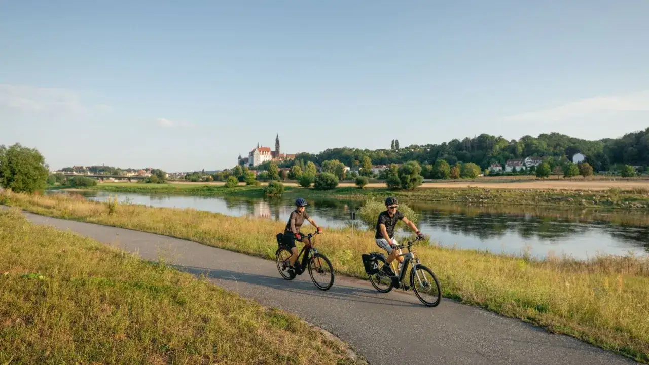 Dwie osoby na rowerach elektrycznych przemierzają malownicze trasy rowerowe Czech, podziwiając rzekę i zamek na wzgórzu.