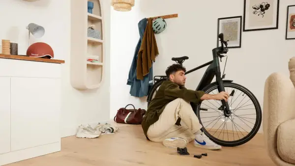 Mężczyzna siedzi na podłodze i majstruje przy rowerze. W tle wieszak z ubraniami i kask, a obok niego **scentrowane koło** roweru.