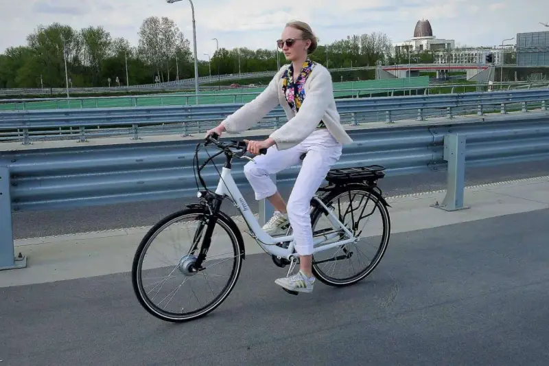 Kobieta w białych spodniach i sweterku jedzie na rowerze elektrycznym. Przepisy dotyczące rowerów elektrycznych są ważne dla bezpieczeństwa.